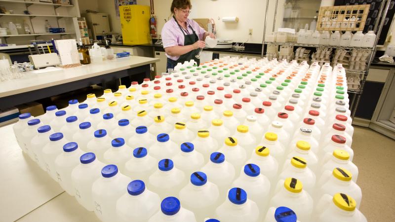 Person pouring liquid into glass tube beside table with various jugs